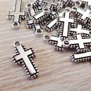 May include: A pile of silver-toned metal cross charms with a beaded border. The charms have a loop at the top for attaching to jewelry or other crafts.