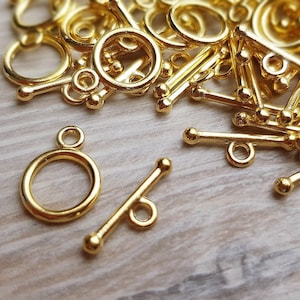 Könnte beinhalten: Nahaufnahme von goldfarbenen Metall-Schiebeverschlüssen. Die Verschlüsse bestehen aus zwei Teilen: einem runden Ring und einer Stange mit einer kleinen Schlaufe an einem Ende. Die Verschlüsse liegen verstreut auf einer Holzoberfläche.