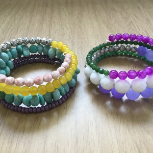 May include: Two bracelets made of colorful beads. The first bracelet is made of purple, white, and green beads. The second bracelet is made of yellow, pink, turquoise, and purple beads.