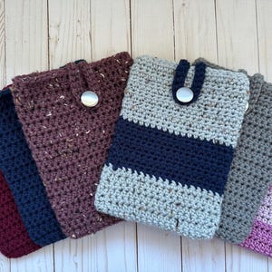 May include: A collection of handmade crochet phone cases in various colors. The cases include burgundy, navy, speckled maroon, gray with a navy stripe, gray, and purple. Each case has a button closure.