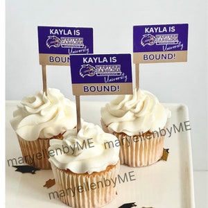 May include: Three white cupcakes with white frosting topped with purple and gold graduation themed cupcake toppers that say "Kayla is Bound!" Western Carolina University Catamounts.