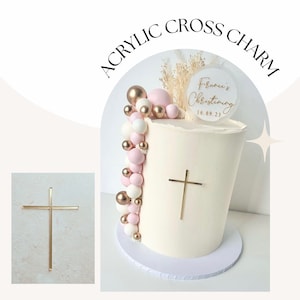 Könnte beinhalten: Ein weißer Kuchen, verziert mit rosa und goldenen Luftballons und einem goldenen Kreuz-Anhänger. Der Kuchen ist mit einem durchsichtigen Acrylschild verziert, auf dem "Francie's Christening 16.09.23" steht.