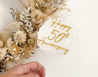May include: A white acrylic cake topper with a gold inscription that reads "Happy 50th Anniversary".