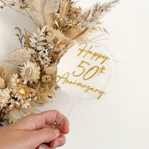 May include: A white acrylic cake topper with a gold inscription that reads "Happy 50th Anniversary".
