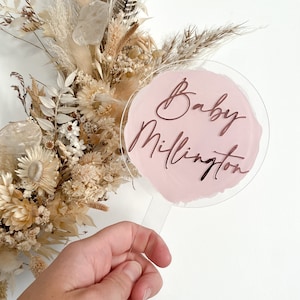 May include: A clear acrylic cake topper with a pink circle and the text "Baby Millington" written in rose gold script.
