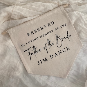 K&ouml;nnte beinhalten: Ein beigefarbenes Canvas-Banner mit schwarzem Text, der "RESERVED IN LOVING MEMORY OF THE Father of the Bride JIM DANCE" lautet.