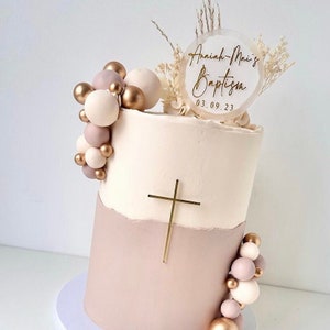 May include: A two-tiered cake decorated with a gold cross, a garland of beige and gold balloons, and a clear acrylic topper with the text "Anahlah-Mai's Baptism 03.09.23".