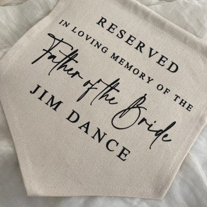 K&ouml;nnte beinhalten: Ein wei&szlig;es Canvas-Banner mit schwarzem Text, der "Reserviert in liebevoller Erinnerung an den Vater der Braut Jim Dance" lautet.