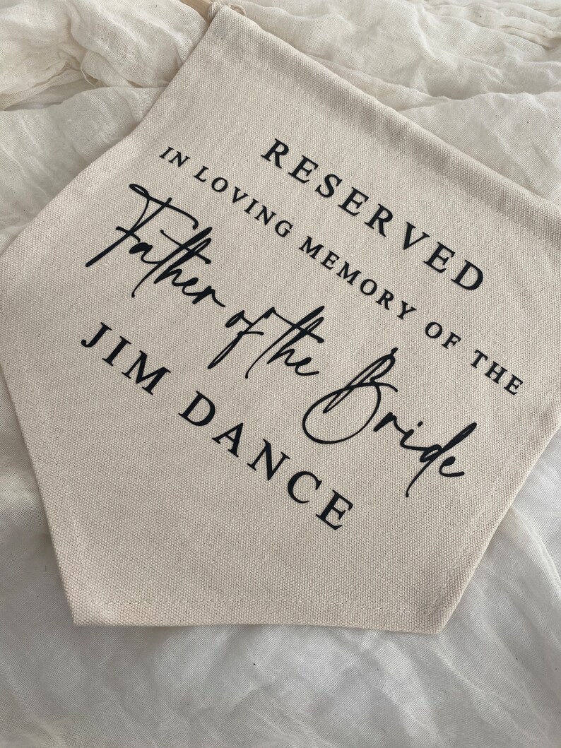 K&ouml;nnte beinhalten: Ein wei&szlig;es Canvas-Banner mit schwarzem Text, der "Reserviert in liebevoller Erinnerung an den Vater der Braut Jim Dance" lautet.