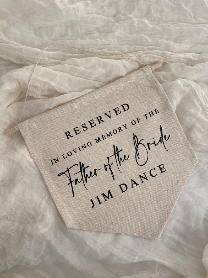 K&ouml;nnte beinhalten: Ein beigefarbenes Canvas-Banner mit schwarzem Text, der "RESERVED IN LOVING MEMORY OF THE Father of the Bride JIM DANCE" lautet.