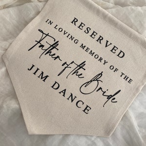 K&ouml;nnte beinhalten: Ein wei&szlig;es Canvas-Banner mit schwarzem Text, der "Reserviert in liebevoller Erinnerung an den Vater der Braut Jim Dance" lautet.