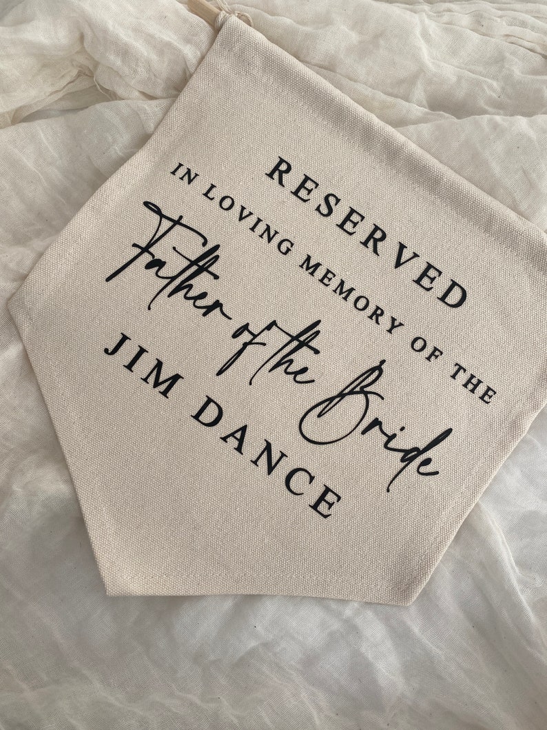 K&ouml;nnte beinhalten: Ein wei&szlig;es Canvas-Banner mit schwarzem Text, der "Reserviert in liebevoller Erinnerung an den Vater der Braut Jim Dance" lautet.