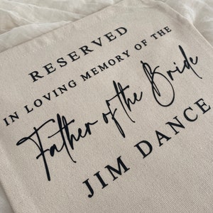 K&ouml;nnte beinhalten: Eine beige Canvas-Tragetasche mit schwarzem Text, der "Reserviert in liebevoller Erinnerung an den Vater der Braut Jim Dance" lautet.
