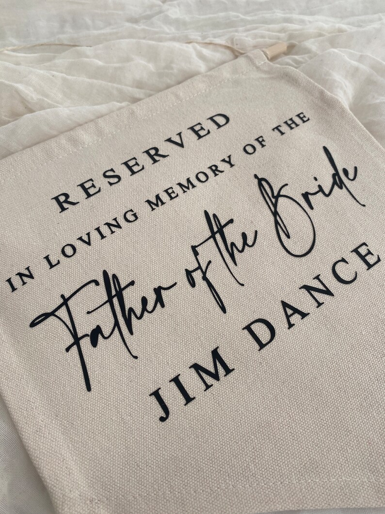 K&ouml;nnte beinhalten: Eine beige Canvas-Tragetasche mit schwarzem Text, der "Reserviert in liebevoller Erinnerung an den Vater der Braut Jim Dance" lautet.