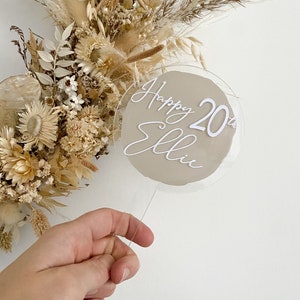 Könnte beinhalten: Ein runder, transparenter Acryl-Kuchentopper mit beigefarbenem Hintergrund und weißer Beschriftung, die "Happy 20th Ellie" lautet.