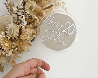 May include: A round, clear acrylic cake topper with a beige background and white lettering that says "Happy 20th Ellie".