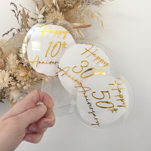 May include: Three clear acrylic round cake toppers with gold lettering. The toppers say "Happy 10th Anniversary", "Happy 30th Anniversary", and "Happy 50th Anniversary".