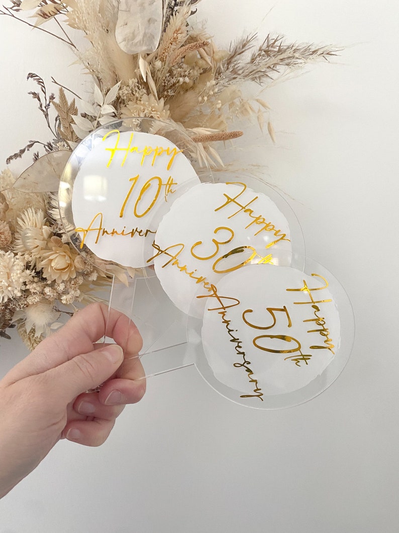 May include: Three clear acrylic round cake toppers with gold lettering. The toppers say "Happy 10th Anniversary", "Happy 30th Anniversary", and "Happy 50th Anniversary".