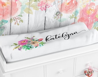 May include: A white baby changing mat featuring a floral design in pink, purple, and green. The name "Ratelynn" is written in black script. The mat rests on a white dresser, with floral wallpaper behind.