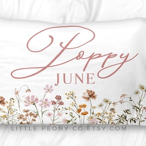 May include: White pillowcase with the name "Poppy" and the month "June" in a soft pink script. The bottom edge features a watercolor floral design with wildflowers in shades of pink, orange, and brown.
