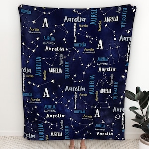 Pode incluir: Uma manta azul e branca com um padrão de constelação e o nome "Aurelia" repetido por toda parte. A manta mede 127 cm por 152 cm.