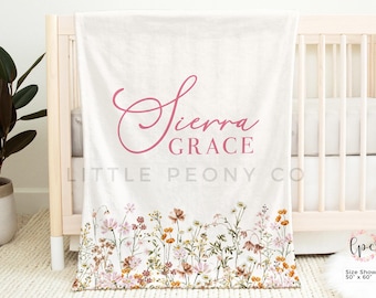 May include: A white baby blanket with a floral border and the name "Sierra Grace" in pink script. The blanket is draped over a cot. The bottom edge features a colorful wildflower design. The blanket measures 127cm x 152cm.