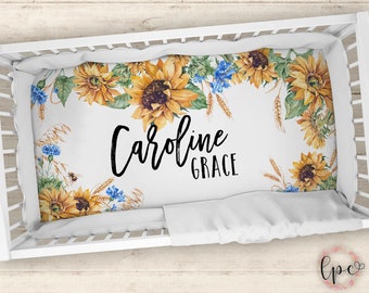 Puede incluir: Sábana de cuna blanca con un borde de girasoles y trigo. La sábana tiene el nombre "Caroline Grace" escrito en cursiva negra.