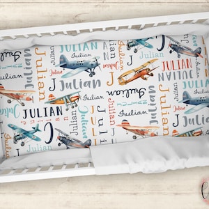 May include: A white crib sheet with a blue and orange airplane pattern. The sheet is covered in the name "Julian" repeated many times.