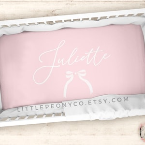 May include: A baby crib with a pink fitted sheet. The sheet has the name "Juliette" in white script, along with a bow graphic. The text "LITTLEPEONYCO.ETSY.COM" is printed at the bottom of the sheet.