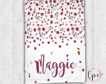 May include: White blanket featuring a floral design in pink, orange, and purple hues. The name "Maggie" is displayed in a cursive font. The blanket's design is complemented by a soft, inviting texture.