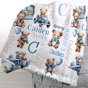 Puede incluir: Una manta de bebé blanca con ilustraciones de osos de peluche, aviones y globos en tonos azules y marrones. La manta está personalizada con los nombres "Caiden" y "Avery".