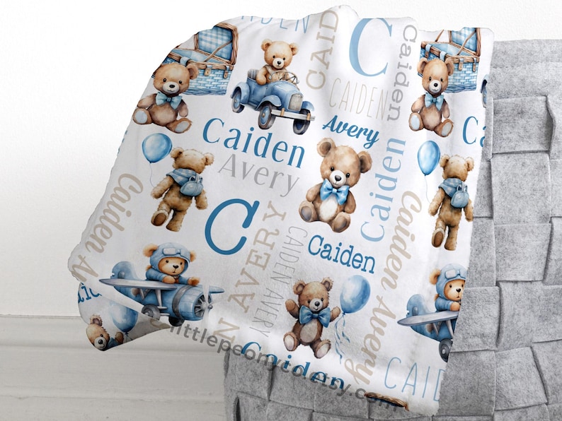 May include: A white baby blanket featuring watercolor illustrations of teddy bears, airplanes, and balloons in shades of blue and brown. The blanket is personalized with the names "Caiden" and "Avery".