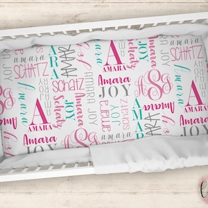 May include: White crib sheet with a pink and teal pattern featuring the names "Amara" and "Joy" repeated throughout. The sheet has a white border.