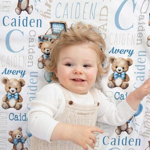 May include: A baby lies on a white blanket decorated with teddy bears, blue balloons, and the names "Caiden" and "Avery" in blue. The blanket has a whimsical, nursery theme.
