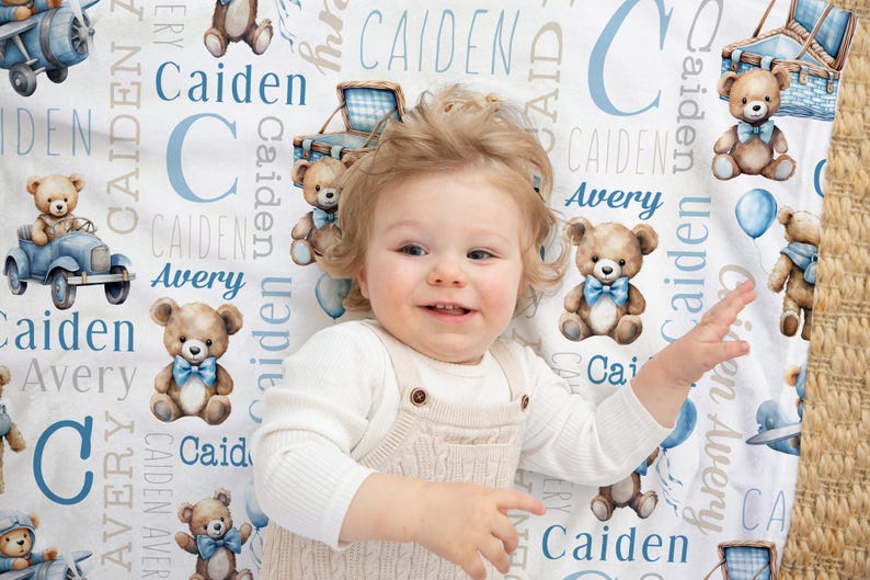 May include: A baby lies on a white blanket decorated with teddy bears, blue balloons, and the names "Caiden" and "Avery" in blue. The blanket has a whimsical, nursery theme.