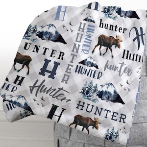 May include: A white and grey plaid blanket with the name "Hunter" in various fonts and colours. The blanket features illustrations of moose, mountains, and trees. The blanket is folded over a grey basket.