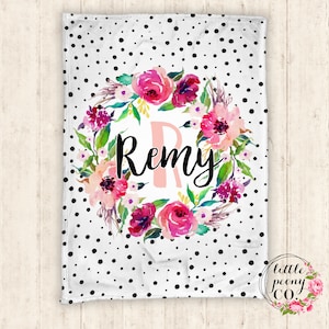 May include: A white fleece blanket with a black polka dot pattern. A pink floral wreath with the name "Remy" in black script is in the center of the blanket.