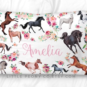 May include: White pillowcase featuring watercolor illustrations of horses in various colors, surrounded by pink and peach floral arrangements. The name "Amelia" is written in pink script.
