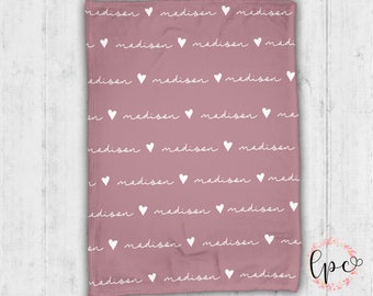 May include: A dusty rose-colored blanket with a repeating pattern of the name "madison" in a white script font, alternating with white heart symbols. The blanket is folded and displayed against a light wood background.