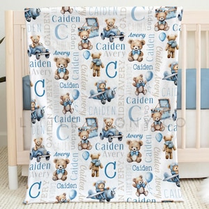 May include: A white baby blanket with a repeating pattern of teddy bears, airplanes, cars, and the names "Caiden" and "Avery" in blue. The blanket is displayed on a crib and measures 50 x 60 inches.