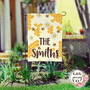 May include: A white garden flag with a honeycomb pattern, bees, and daisies. The text "THE Smiths" is printed in brown on the flag.