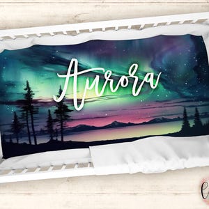 May include: A white crib with a white and green aurora borealis themed crib sheet. The sheet has the name "Aurora" written in white cursive lettering.