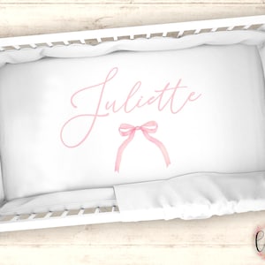 以下が含まれることがあります： ピンクのリボンと「Juliette」という名前が筆記体で書かれた白いベビーベッドシーツです。