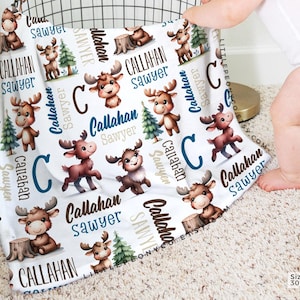 May include: A white baby blanket with a repeating pattern of cartoon moose, trees, and the name "Callahan Sawyer". The blanket measures 30 x 40 inches.