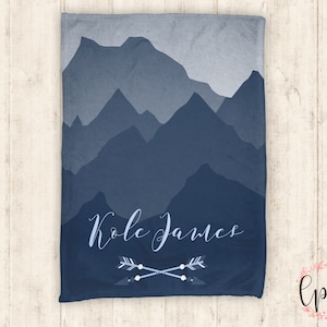 May include: A blue and gray mountain range design on a soft fleece blanket with the name "Kole James" and two crossed arrows in white.