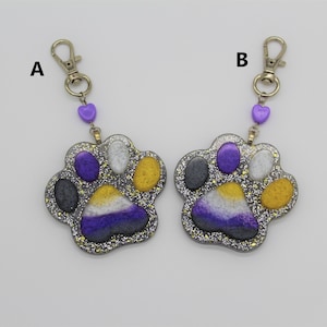 May include: Two silver keychains with purple heart charms and a paw print charm with glitter and the colours of the non-binary pride flag.