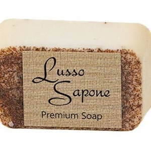 May include: A rectangular bar of off-white soap with a textured, brown, granular surface on the sides. A tan, fabric-textured label is wrapped around the center, featuring the black text Lusso Sapone and Premium Soap in a serif font.