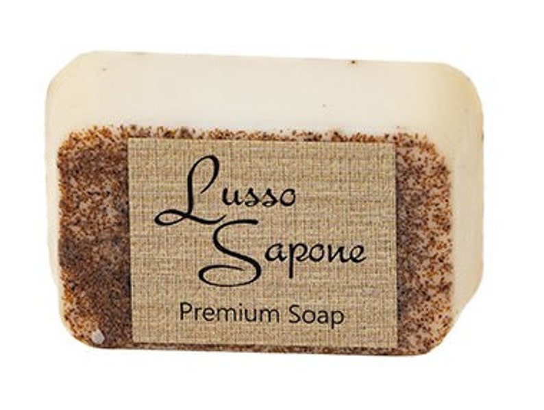May include: A rectangular bar of off-white soap with a textured, brown, granular surface on the sides. A tan, fabric-textured label is wrapped around the center, featuring the black text Lusso Sapone and Premium Soap in a serif font.