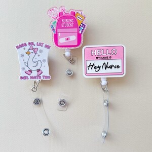 Peut inclure: Trois porte-badges roses et blancs avec différents motifs. L'un représente un sac à dos avec le texte "NURSING STUDENT". Un autre montre une oie avec "HANG ON, LET ME GIRL MATH THIS". Le troisième dit "HELLO MY NAME IS Hey Nurse".