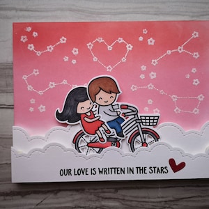 Könnte beinhalten: Handgefertigte Grußkarte mit rosa-rotem Farbverlauf, die ein Paar auf einem Fahrrad zeigt. Die Karte enthält den Text "OUR LOVE IS WRITTEN IN THE STARS" und ein Herz.